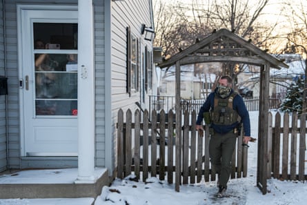 a masked man in tactical gear walks out of a backyard alongside a house in which people are standing inside the front door recording with their phones