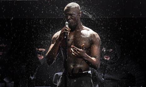 Man with microphone gestures as rain falls; behind him are masked faces in the darkness