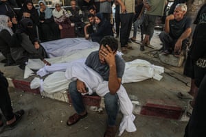 A man sits holding a small body wrapped in a white sheet