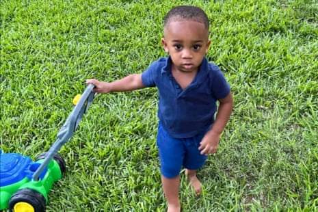A toddler in a blue polo shirt stands barefoot on grass holding a toy lawnmower