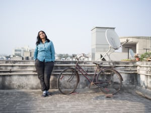 A women leans against a railing next to a bike with a cityscape behind her.