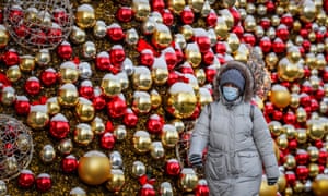 A woman walks on a street in Moscow, Russia. She wears a blue face mask and is backed by red and gold baubles.