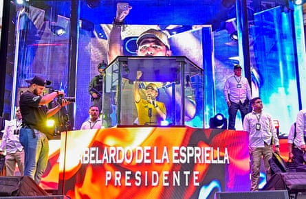 A man gestures from inside a perspex box while surrounding screens show him in closeup.