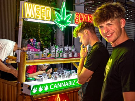 A young man smiles at the camer while another looks at cannabis products displayed on a stall