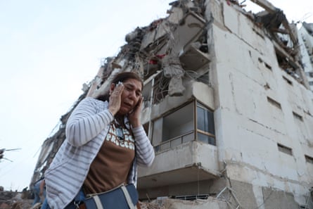A woman holds her hands to her head near a damaged building