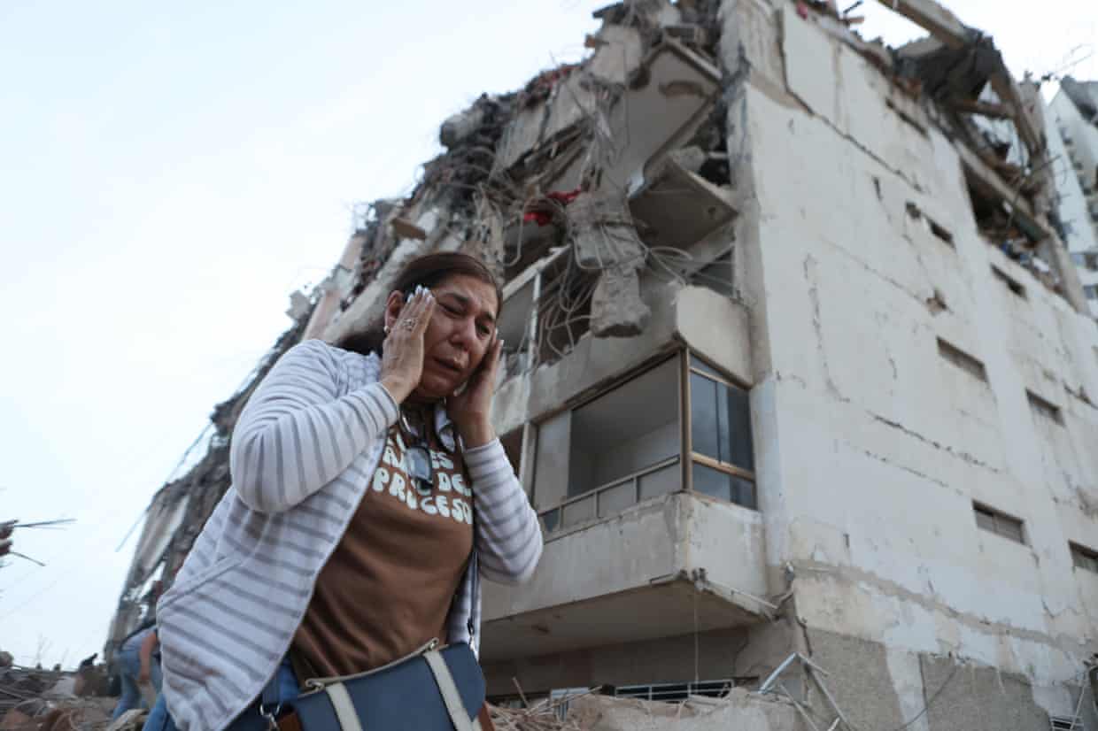 A woman holds her hands to her head near a damaged building