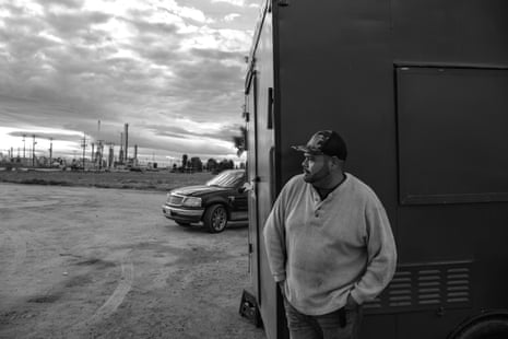 A man in a hat looks to the left, an oil refinery and truck in the background