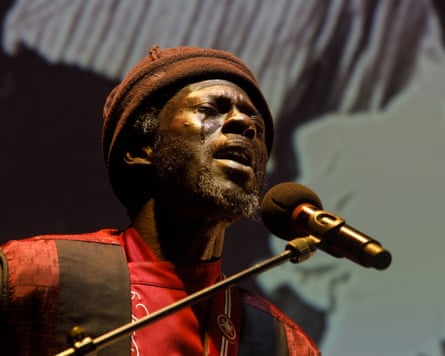 A man in a knitted cap and T-shirt sings in front of a microphone, eyes closed and a tear running down his cheek