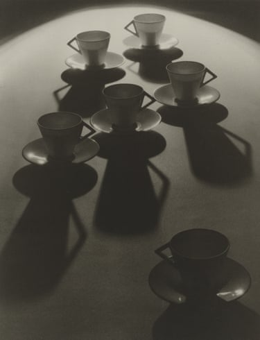 Six teacups and saucers cast long dramatic shadows on a curved surface, in black and white