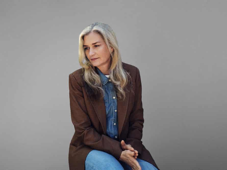A portrait of Marianne Shine today with long, greying hair and wearing a demin shirt, jeans and a brown jacket