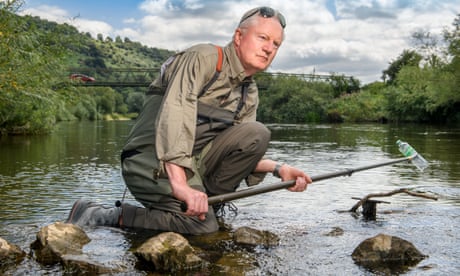 Gordon Green, in fishing overalls and sunglasses pushed onto his forehead, crouches as he takes a plastic bottle out of the River Wye with a stick