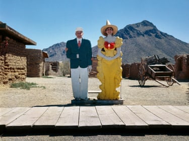 A man and a woman stand, with their heads showing, behind headless models of a man in shirt, tie, blue jacket and white trousers and a woman in a large, flouncy yellow outfit with a big, red bow at the neck