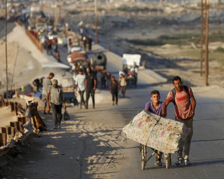 People walk along an asphalt road - a boy next to a man pushes a mattress on a wheelchair,