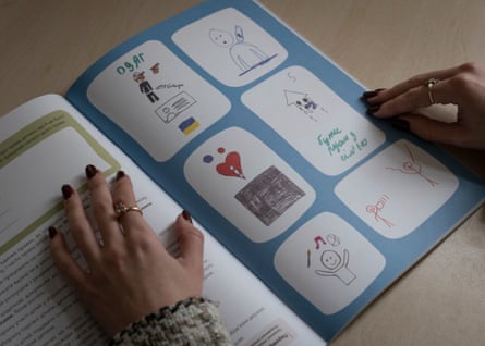 A woman’s hands holding a booklet with Cyrillic writing and a child’s drawings of stick figures, a gun, a Ukrainian flag etc