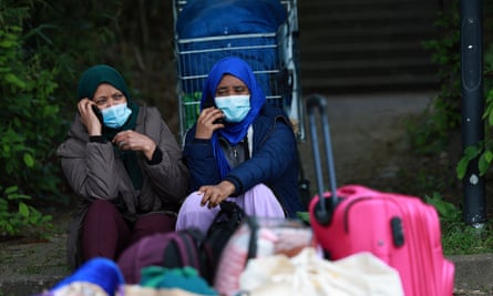 Two women wait with their belongings; they are sitting outside the factory with bags and a large bright pink wheeled suitcase, with a supermarket trolley behind them. They both wear headscarves and blue surgical masks.