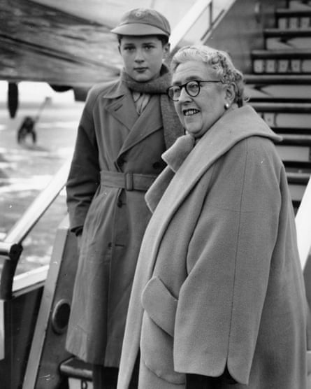 A black and white photo of Agatha Christie standing on the bottom of the steps of a plane in a heavy camel coat, with her grandson, Mathew Prichard, James’s father, wearing a schoolboy cap, belted winter coat, and scarf.