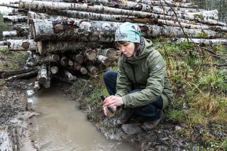 Environmental expert Mari Annala crouches by muddy water near stacked logs, testing a sample