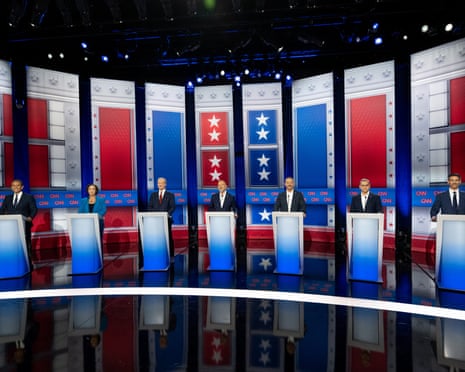 Seven California governor candidates stand behind podiums on a debate stage with red, white, and blue decorations