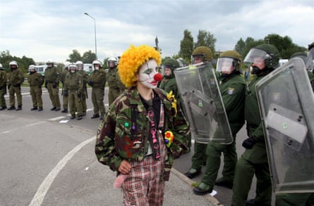 A protester dressed as a clown walks in front of a line of riot police holding shields