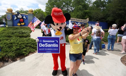 person takes selfie with someone in a mickey mouse costume, with a Trump hat and Ron DeSantis sign