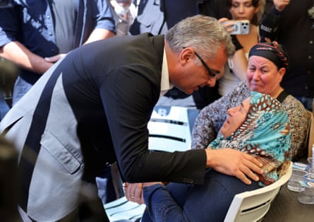 Tufan Erhürman, a grey-haired man wearing glasses and a dark blue jacket, leans over a woman wearing a headscarf who is sitting in a chair with his hand on her shoulder. Other people are sitting by.