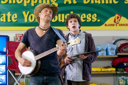 A man wearing a cowboy hat and playing the banjo standing next to a younger man with short curly hair, wearing a blue shirt and a brown hoodie