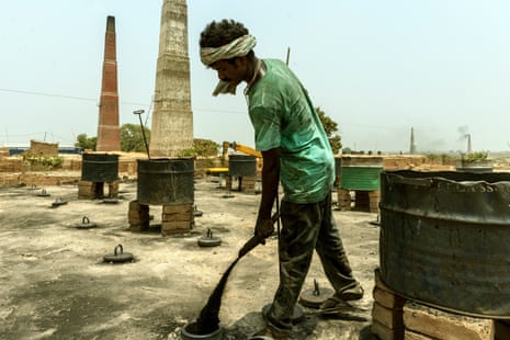 A man wearing cloth around his head and a sweat-soaked shirt lets a scoop of black dust fall into an opening in the kiln.