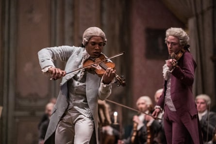 Kelvin Harrison Jr in late 18th-century costume and wig playing a violin with gusto. With orchestra behind him and a second fiddle player to his left