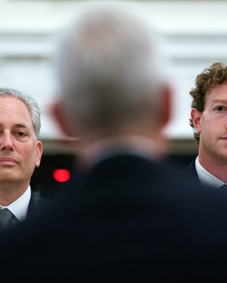 two men in suits sit at dining table, facing another man in suit with back to camera