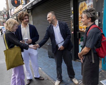 A man, Green party leader Zack Polanski, shakes hand with a woman in a street, while two other people stand close by.
