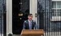 Rishi Sunak issues a statement outside 10 Downing Street: he is standing behind a wooden lectern in front of No 10's black wooden door and black ironwork gateway with a central lamp. It is raining and the building's brickwork looks damp and dark grey behind him.