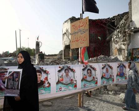 A woman wearing a burqa holds pictures of her children while in the background pictures of other children are displayed against ruins.