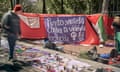 A passerby looks at purple sheets spread out on the pavement covered with a variety of wares, behind which is a red screen strung between two trees covered in hangings with feminist slogans in Spanish and a Palestinian flag