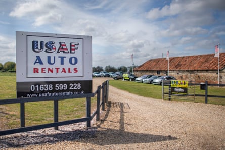 A sign at a gateway reads, in red and blue with stars and stripes, USAF auto rentals. Down the gravel driveway there are cars lined up in front of a barn.