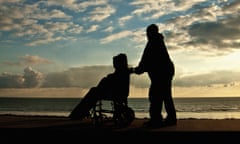 Silhouetted male pushing an anonymous person in a wheelchair along a seafront as the sun sets behind them