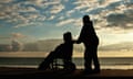 A silhouetted male pushing an unidentifiable person in a wheelchair along seafront as the sun sets.