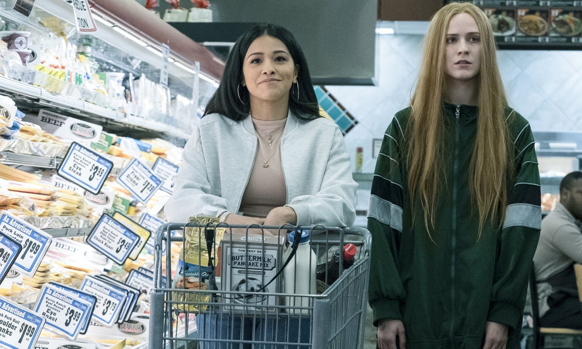 Image is from the film 'Kajillionaire' (2020). Two women stand next to one another in an aisle of a supermarket. One woman is pushing a trolley, wearing a white bomber jacket and hoop earrings. The other has extremely long red hair and is wearing a black jacket.