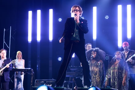 Jarvis Cocker of Pulp, dressed in dark suit and dark shirt, croons into the microphone as his band play behind him