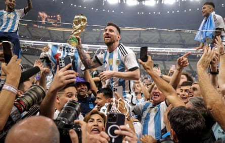 Lionel Messi is held aloft by teammates with the World Cup trophy in his hand as he is surrounded by photographers