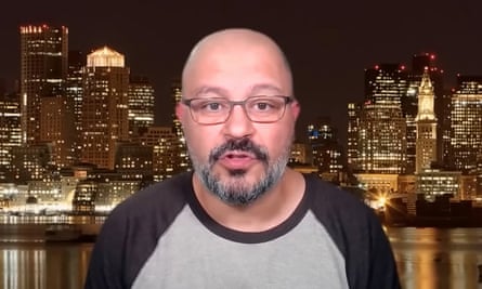 A bald man with a beard and glasses against a skyscrapers lit up at night