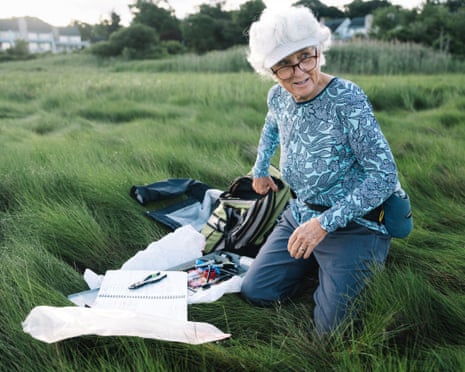A women with grey hair kneels down in a marsh with a notepad, pliers and other items beside her