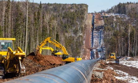A large pipe runs from the bottom left of the picture up a hill lined with tress with diggers on either side of a trench.