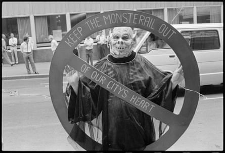 A protestor wearing a skull costume holds a cut-out stop sign with ‘keep the monsterail out of our city’s heart’ written on it
