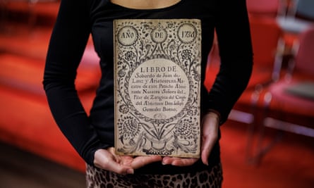 A woman holds up a book with an elaborately illuminated cover with plant and floral motifs in black and white, the title in Spanish, and a date of 1734