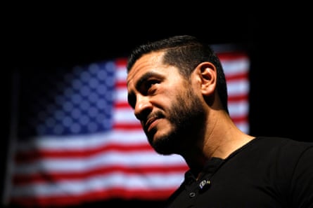 A man with short hair and a beard in front of a projection of an American flag.