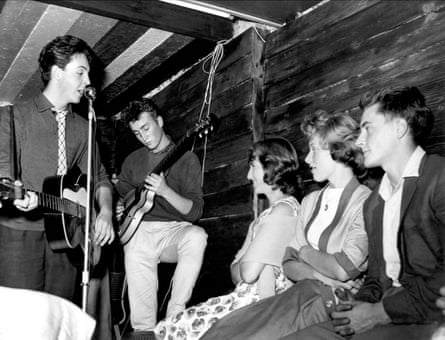 A black and white photo of two men playing the guitar watched by three women on the right