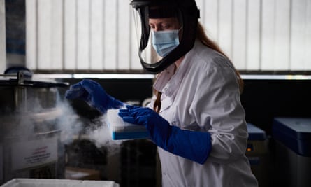 Woman in PPE equipment and large dark blue gloves holds a steaming cup in a lab.