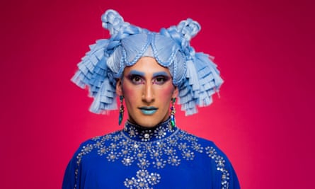 Amrou Al-Kadhi in drag, with bright blue hair in plaits and tied up, on a pink/red background