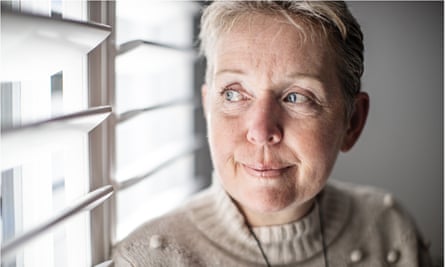 Closeup of Tracy Hickman’s face as she stands by the light in front of a window with white shutters; she is looking outwards and is smiling