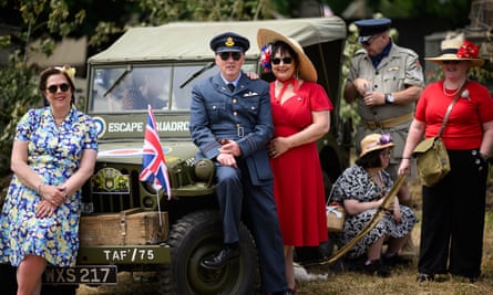 Enthusiasts wearing replica WWII-era attire stand in front of a vintage army vehicle, the men wearing uniform, the women wearing 1940s-era dresses, in a re-enactment camp in Sainte-Mère-Église, north-western France, as part of the D-day commemorations